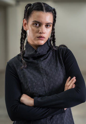A woman wearing a black textured cotton vest with a funnel neckline, over a long-sleeved top, with her arms crossed.