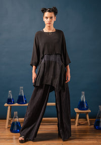 Woman in a black outfit standing in front of blue liquid in beakers on wooden stools against a blue wall.