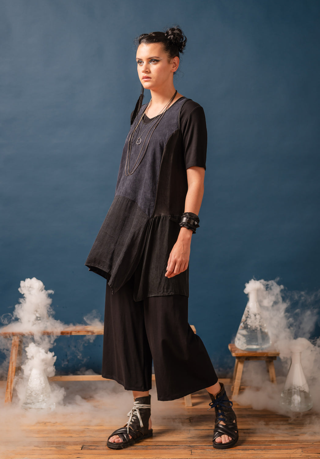 Woman in a black outfit standing in front of blue liquid in beakers on wooden stools against a blue wall.