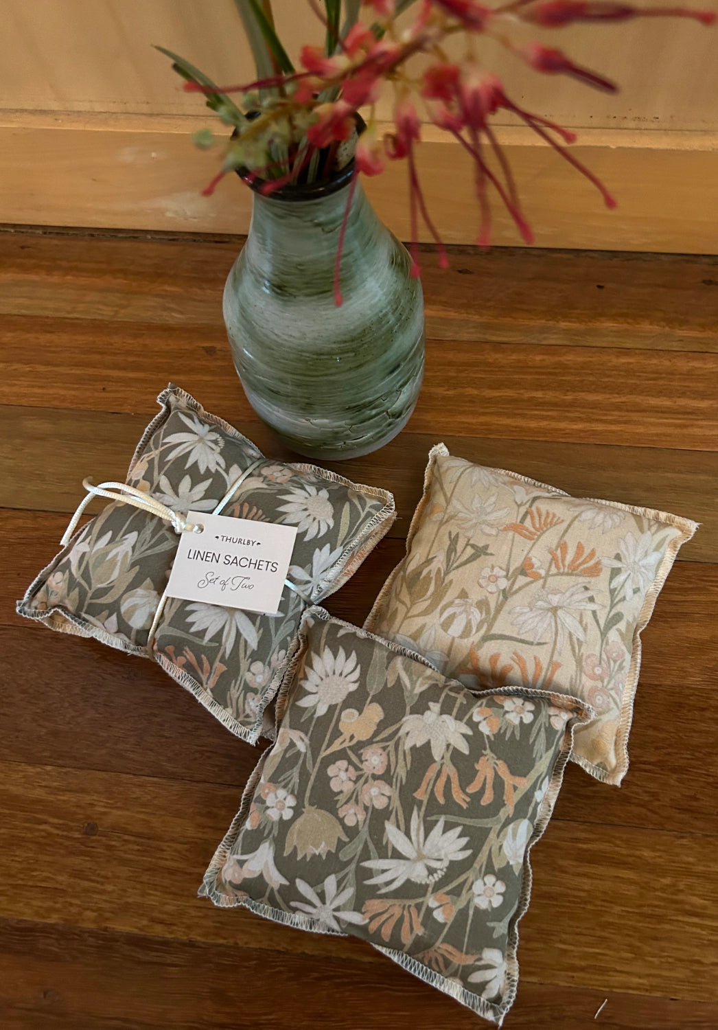 Three floral-patterned linen sachets on a wooden surface with a vase of flowers in the background.