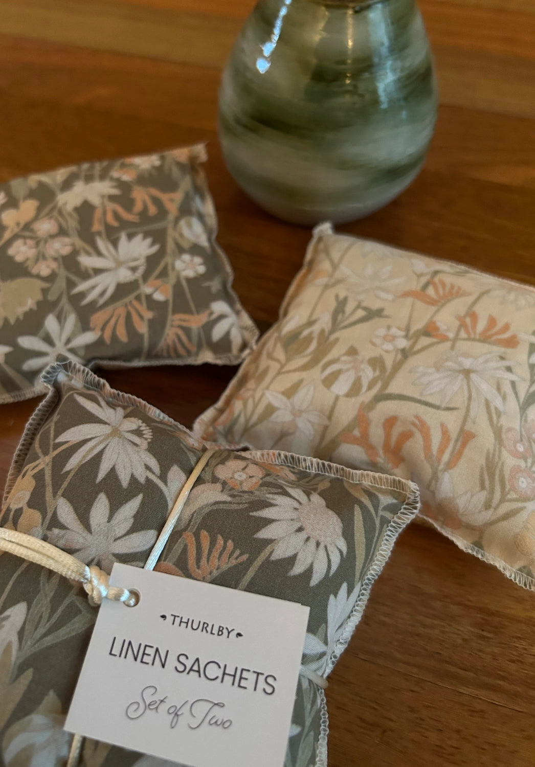 Three floral-patterned linen sachets on a wooden surface with a vase of flowers in the background.
