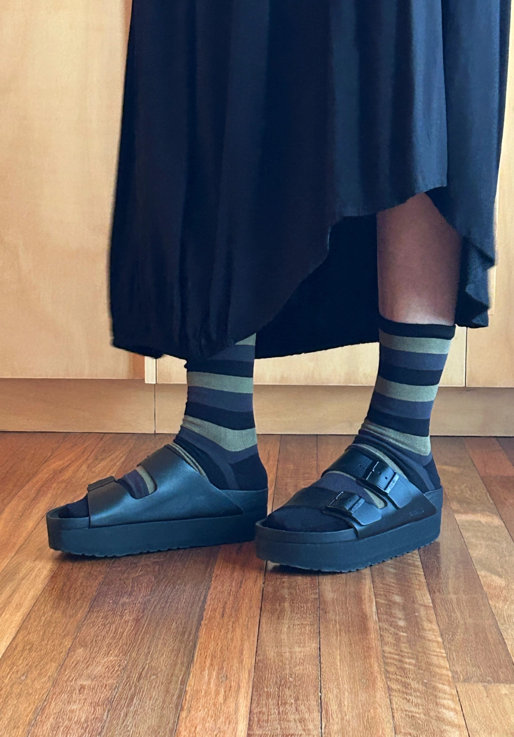 A person wearing dark navy platform slide sandals with wide straps, paired with navy and gray striped crew socks and a dark skirt, standing on a wooden floor.