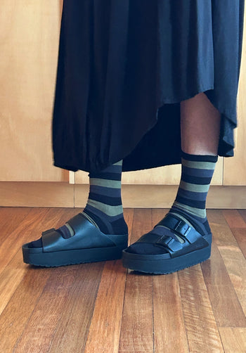 A person wearing dark navy platform slide sandals with wide straps, paired with navy and gray striped crew socks and a dark skirt, standing on a wooden floor.