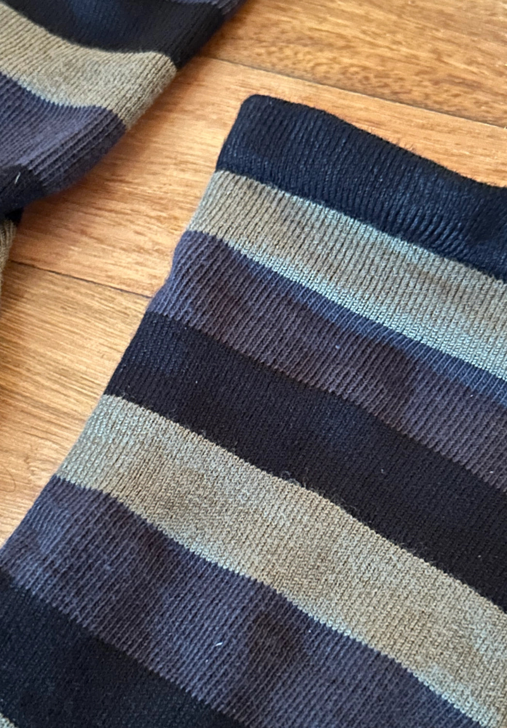 A close-up of a striped knit sock laid flat on a wooden floor, showing horizontal bands of dark navy, charcoal gray, and muted sage green.
