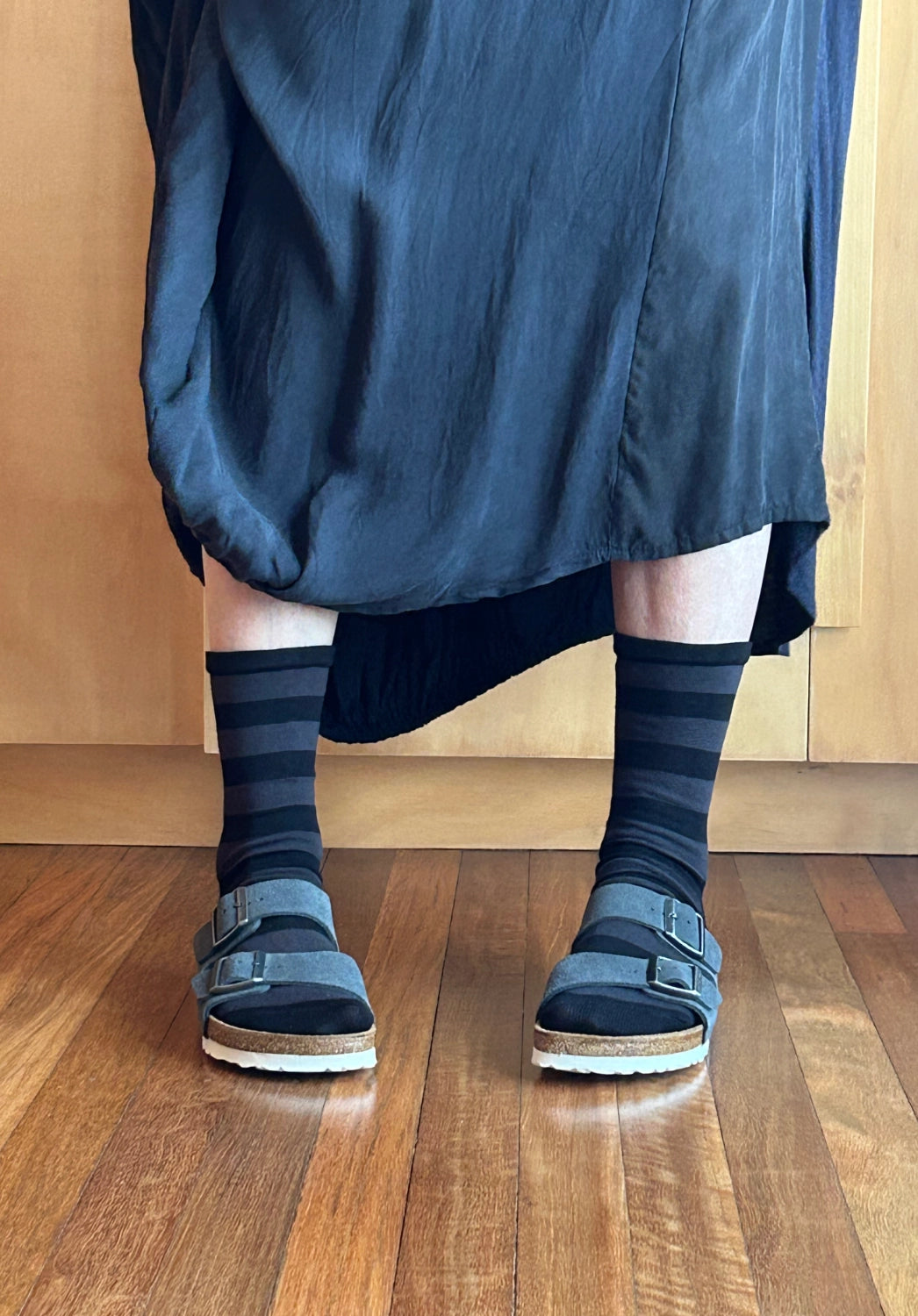 A person wearing dark navy and black striped crew socks with dark blue suede slide sandals and a dark blue skirt, standing on a wooden floor.