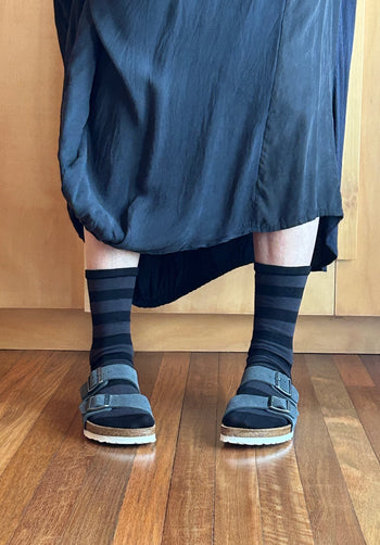 A person wearing dark navy and black striped crew socks with dark blue suede slide sandals and a dark blue skirt, standing on a wooden floor.