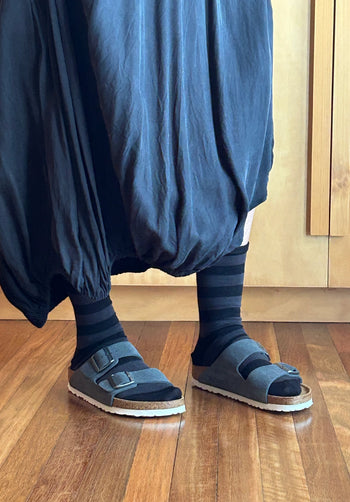 A person wearing a dark blue baggy skirt, dark striped socks, and dark gray slide sandals with cork soles, standing on a wooden floor.