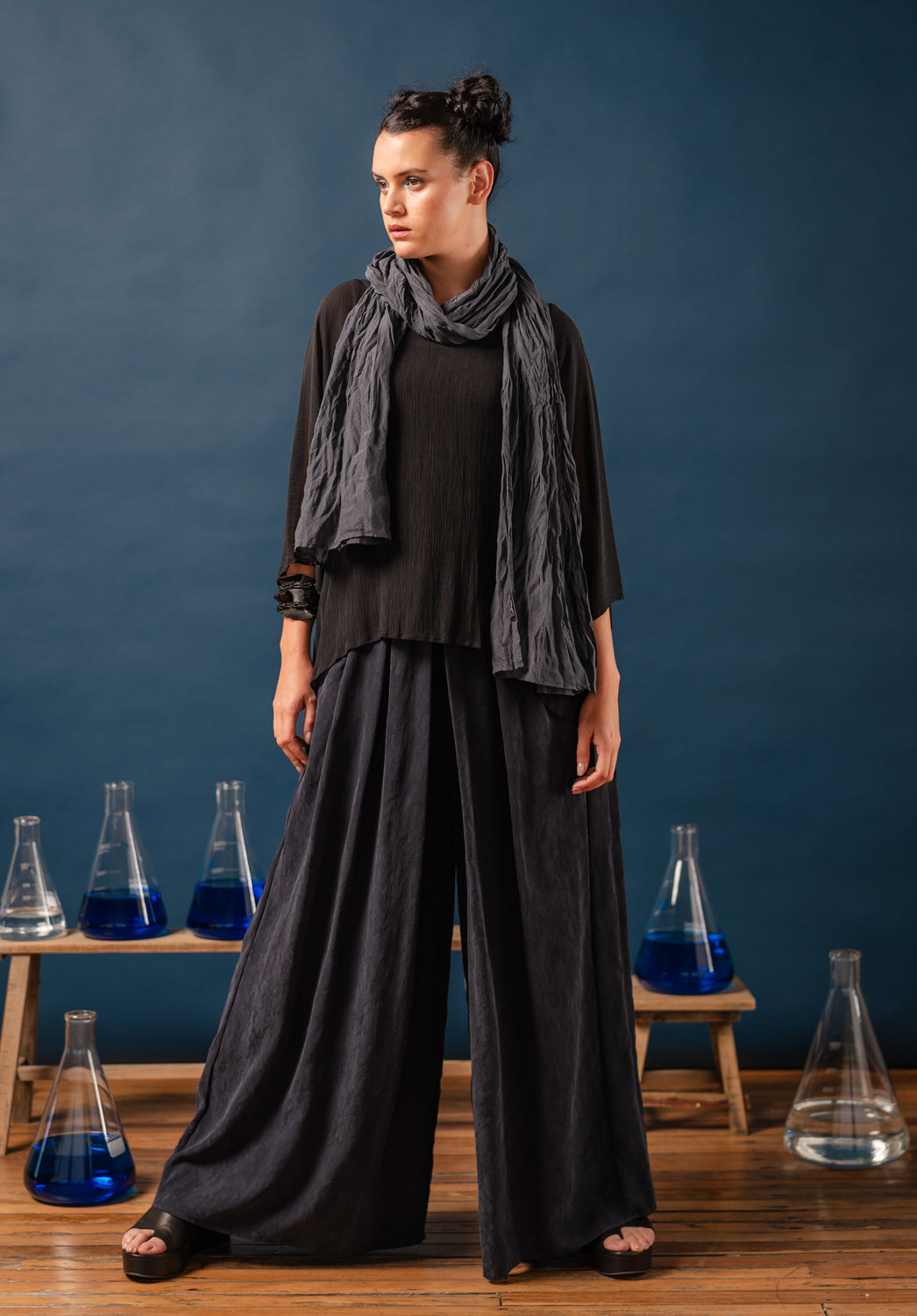 Woman in a black with ink cupro pants and a gray scarf standing in front of blue beakers filled with a blue liquid on a wooden shelf.